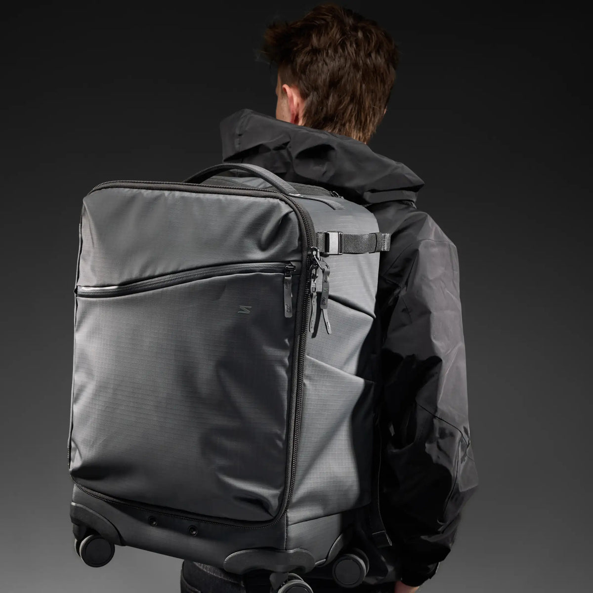 A man viewed from behind wearing a large dark gray rolling backpack-style duffel bag over his shoulders, against a dark studio background.