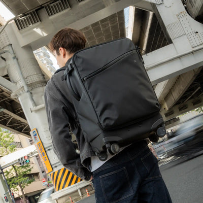 A man wearing a dark gray backpack with a built-in rolling suitcase base, photographed from behind in an urban street setting under an elevated train track.